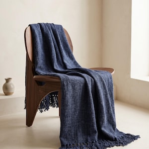 May include: A dark blue woven throw blanket with a textured finish and fringed edges draped over a curved wooden chair. The fabric features a visible cross-hatch pattern throughout, and the fringe hangs from the bottom edge of the blanket.