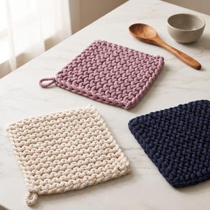 May include: Three square, thick, hand-knitted potholders in mauve, cream, and navy blue. Each potholder features a dense, textured weave pattern and a small loop attached to one corner for hanging. The items are arranged on a flat surface.