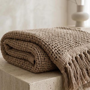 Może przedstawiać: Złożony, beżowy dzianinowy koc z frędzlami. Koc ma wzór o otwartym splocie. Frędzle znajdują się na dolnej krawędzi. Koc spoczywa na jasnej, kwadratowej powierzchni. Koc jest w neutralnym kolorze.