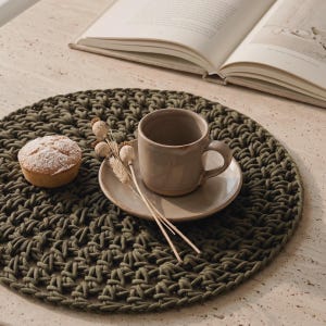 Pode incluir: Um lugar americano redondo de crochê em verde oliva escuro com uma textura de cordão grosso e entrelaçado. Sobre o lugar americano, há uma xícara e pires de cerâmica bege claro, um pequeno doce redondo polvilhado e três papoulas secas.