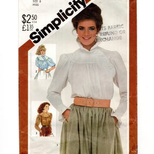 May include: A Simplicity 5575 sewing pattern envelope for a miss size 8. The cover features a white long-sleeved blouse with a high collar and a light green skirt cinched with a wide tan belt. Two smaller illustrations show variations in blue and brown.