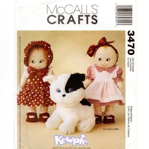 May include: McCall's Crafts doll pattern featuring two Kewpie dolls in dresses and a plush black and white dog. The dolls have painted faces and are wearing bonnets and bows. The pattern number is 3470.