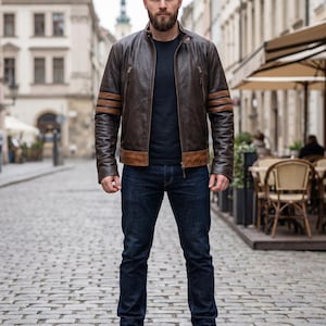 Può includere: Una giacca in pelle marrone scuro con colletto alla coreana, due tasche verticali con zip sul petto e due strisce orizzontali in pelle color cuoio su ogni manica, indossata sopra una t-shirt nera liscia con jeans blu scuro e stivali in pelle marrone.