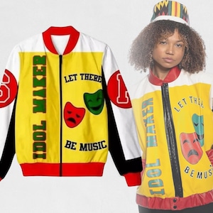 May include: A yellow, white, and black bomber jacket with the text "IDOL MAKER" and "LET THERE BE MUSIC". The jacket features red and green theater masks and the number "1" and "3". The jacket is worn by a person wearing a matching hat.