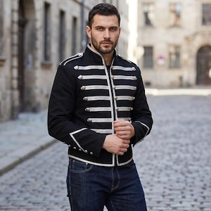 May include: A black military-style jacket featuring a high collar, long sleeves, and silver-toned decorative buttons arranged in horizontal rows across the front. The jacket includes silver piping trim along the edges, cuffs, and shoulder epaulets.