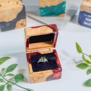 May include: A wooden ring box with a red resin inlay, open to reveal a silver ring with a green gemstone. The box is lined with black velvet. Other wooden blocks with resin are in the background.