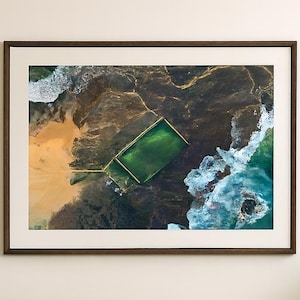 May include: A framed aerial photograph of a green rectangular pool surrounded by rocks and ocean waves. The artwork is displayed above a beige sofa with a green cushion, a round wicker coffee table, and a white lamp.