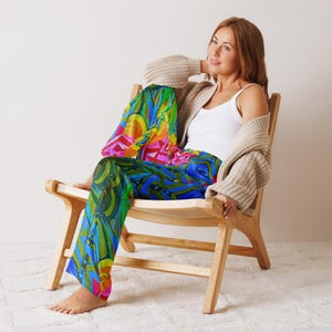 May include: Abstract print pajama trousers in blue, green, yellow, pink, and red. A person is wearing the trousers while sitting in a wooden chair. They are also wearing a white vest top and a beige cardigan.