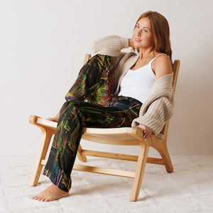May include: A person wearing black pajama trousers with a vibrant abstract pattern, a white vest top, and a beige cardigan, seated in a light wood chair. The trousers have green, yellow, and red colours.