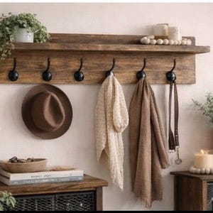 May include: A rustic wooden wall-mounted shelf with six black hooks, holding a brown hat, two scarves, and a leather belt. The shelf also displays a potted plant and decorative items, creating a cozy, organized space.