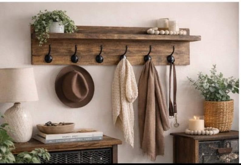 May include: A rustic wooden wall-mounted shelf with six black hooks, holding a brown hat, two scarves, and a leather belt. The shelf also displays a potted plant and decorative items, creating a cozy, organized space.