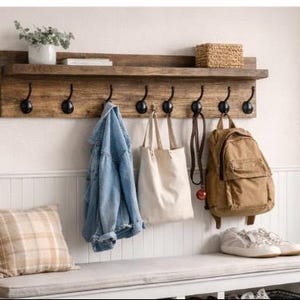 May include: A rustic wooden wall shelf with black hooks, holding a denim jacket, canvas tote bag, and a brown backpack. A bench below holds sneakers and a plaid pillow. A woven basket and a plant in a white pot are also visible.