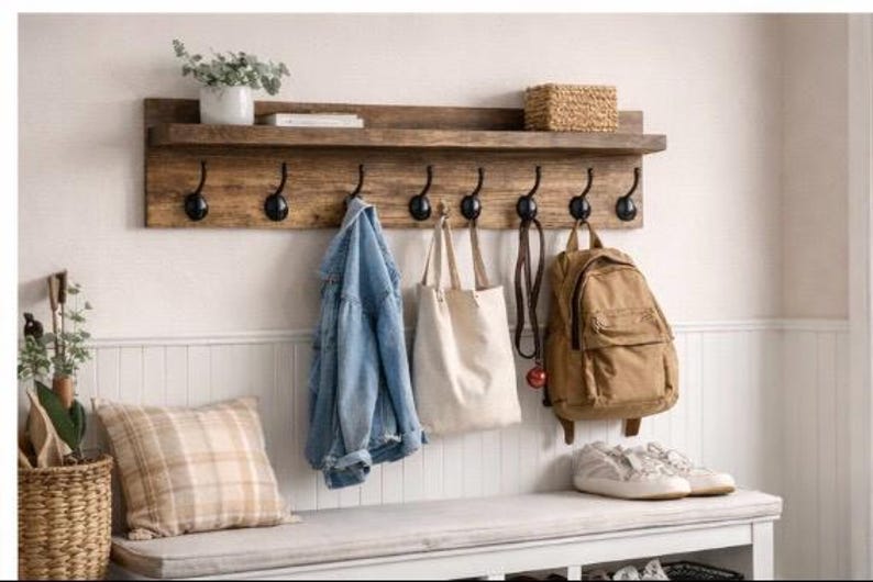 May include: A rustic wooden wall shelf with black hooks, holding a denim jacket, canvas tote bag, and a brown backpack. A bench below holds sneakers and a plaid pillow. A woven basket and a plant in a white pot are also visible.