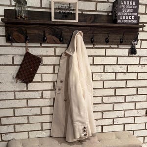 May include: A dark stained wooden shelf with hooks, holding a cream-colored coat and a brown purse. The shelf also displays a framed sign that says "I love you more than bacon" and a black box with inspirational text. A bench sits below.