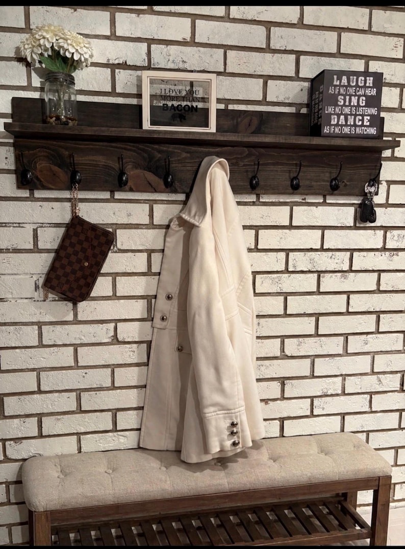 May include: A dark stained wooden shelf with hooks, holding a cream-colored coat and a brown purse. The shelf also displays a framed sign that says "I love you more than bacon" and a black box with inspirational text. A bench sits below.