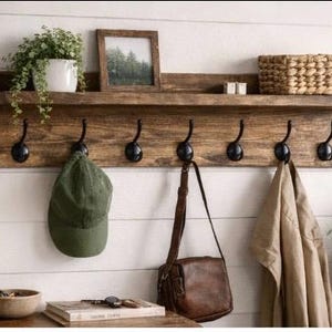 May include: A rustic wooden wall shelf with six black metal hooks. A green cap, brown leather bag, and tan jacket hang from the hooks. The shelf holds a framed picture, a woven basket, and a potted plant.