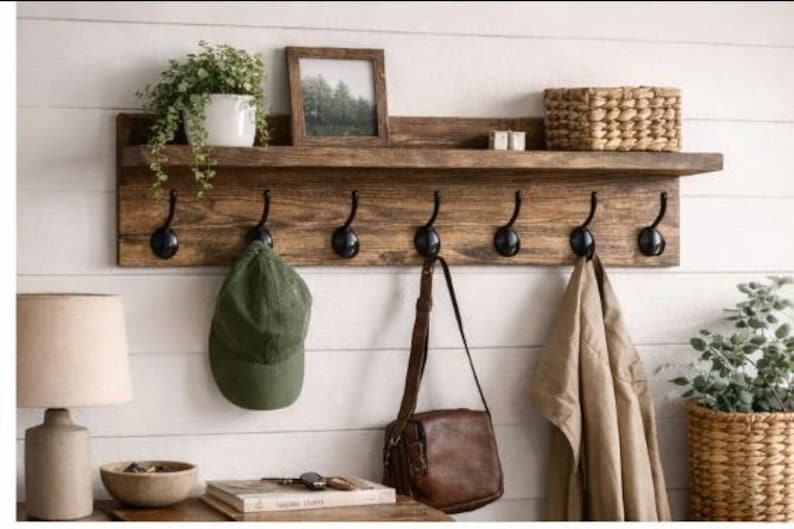 May include: A rustic wooden wall shelf with six black metal hooks. A green cap, brown leather bag, and tan jacket hang from the hooks. The shelf holds a framed picture, a woven basket, and a potted plant.