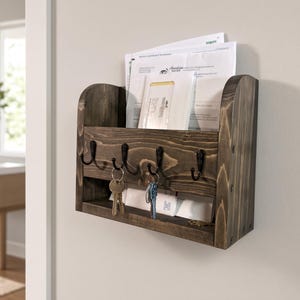 May include: A dark brown stained wooden wall-mounted organizer featuring a top slot for mail and documents, and a lower section equipped with four black metal hooks. Two sets of metal keys are hanging from the hooks, and white envelopes are visible.