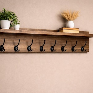 May include: A rustic wooden wall shelf with eight black metal hooks. The shelf is stained a warm brown and has a small ledge. Two white potted plants and a vase with dried wheat are on the shelf.