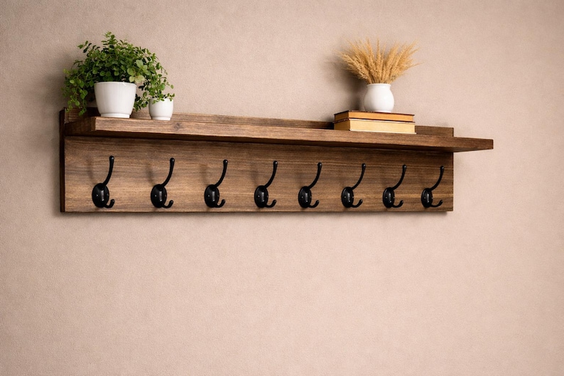 May include: A rustic wooden wall shelf with eight black metal hooks. The shelf is stained a warm brown and has a small ledge. Two white potted plants and a vase with dried wheat are on the shelf.