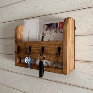 May include: A wooden mail and key holder with a dark stain finish. The organizer has a slot for mail and four black metal hooks for keys. The holder is mounted on a light wood-paneled wall. The mail slot contains envelopes and a card that says "It's a Bridal".
