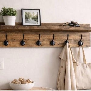May include: A rustic wooden wall shelf with six black metal hooks. A small potted plant, framed picture, and keys rest on the shelf. A tan coat and tote bag hang from the hooks. The shelf is mounted on a white wall.