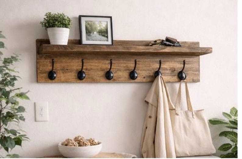 May include: A rustic wooden wall shelf with six black metal hooks. A small potted plant, framed picture, and keys rest on the shelf. A tan coat and tote bag hang from the hooks. The shelf is mounted on a white wall.