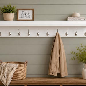 May include: A white wall-mounted coat rack with ten silver metal hooks. The shelf holds a potted plant, a framed sign reading "home is where we hang our coats", and two stacked books. A tan jacket hangs from one hook above a wooden bench.