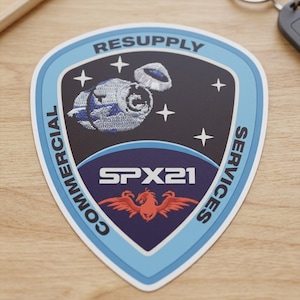 May include: A shield-shaped sticker with a space-themed design. The sticker features a blue border, the words "RESUPPLY" and "COMMERCIAL SERVICES", and the text "SPX21". A spacecraft and stars are also depicted.