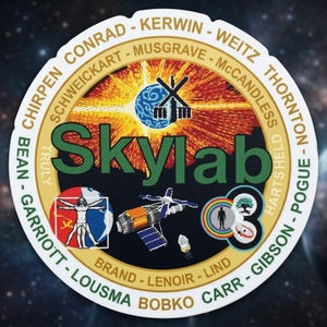 May include: A circular Skylab mission patch with a black background and a gold border. The patch features the word "Skylab" in green, surrounded by names and various illustrations, including a satellite and a human figure.
