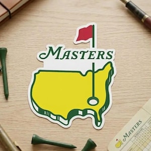 May include: A Masters golf tournament sticker featuring a yellow map outline with a green border, a red flag, and the word "Masters" in green script. The sticker is on a light wood surface, with golf tees and a scorecard nearby.