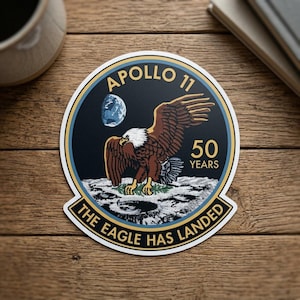May include: A circular sticker featuring the Apollo 11 mission emblem. The design includes a bald eagle landing on the moon, with the Earth in the background. Text reads "Apollo 11" and "The Eagle Has Landed", celebrating 50 years.