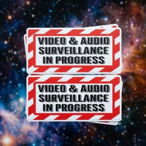 May include: Two rectangular stickers featuring a white background with a red and white diagonal striped border. The text in black, bold, sans-serif font reads: VIDEO & AUDIO SURVEILLANCE IN PROGRESS. The stickers are stacked vertically.