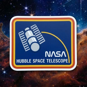 May include: A rectangular sticker featuring the NASA logo and the Hubble Space Telescope. The design includes a blue background with a white and silver illustration of the telescope, a yellow arc, and the NASA text in white. The border is red, yellow, and white.