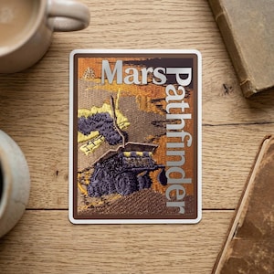May include: Embroidered patch featuring the words "Mars Pathfinder" in silver lettering. The patch depicts a Mars rover in shades of purple, yellow, and brown against a textured background. The patch has a brown border and a white backing.