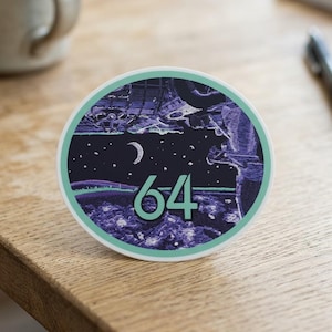 May include: A round sticker with a dark blue and purple illustration of a night sky with a crescent moon and stars. The number "64" is in the center in a teal color, surrounded by a teal border.