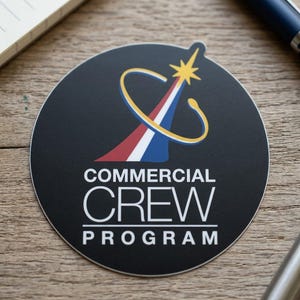 May include: A round black sticker featuring the Commercial Crew Program logo. The logo includes a stylized star, a curved yellow and blue design, and a red, white, and blue arrow. The words "COMMERCIAL CREW PROGRAM" are printed in white.