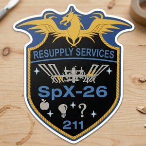 May include: A sticker featuring a shield design with a gold phoenix and the text "RESUPPLY SERVICES". The shield is black with the text "SpX-26" and other symbols. The number "211" is at the bottom.
