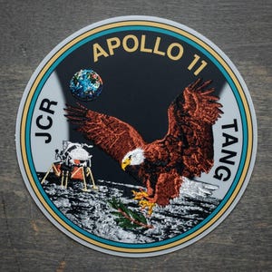 May include: Circular patch featuring the Apollo 11 mission logo. The design includes a brown eagle with outstretched wings, a depiction of the Earth, and a lunar module on a gray surface. The text "APOLLO 11" and other text are in gold.