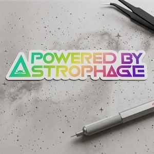 May include: A colorful sticker with the text "POWERED BY ASTROPHAGE" in a rainbow gradient. The sticker has a triangular design with a spaceship graphic. Tweezers and a silver pen are in the background.