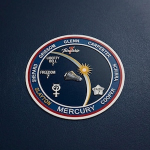 May include: A circular patch with a blue background and a red and white border. The patch features the word "MERCURY" and the names of astronauts. It includes a stylized sun, a rocket, and the text "Friendship 7".