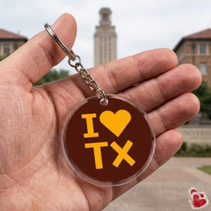 May include: A clear acrylic keychain with a brown background and the words "I ♥ TX" in yellow. The keychain is attached to a silver chain and ring. The background is blurred, showing a building.