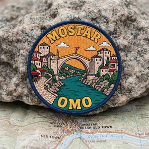 May include: A circular embroidered patch featuring a landscape of Mostar with the Stari Most bridge over a teal river. The sky is orange with white clouds. The text MOSTAR is arched at the top and OMO is at the bottom in yellow letters.
