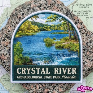 May include: A semi-circular embroidered patch featuring a detailed landscape of a blue river with a small waterfall, surrounded by green trees and foliage. The bottom dark green border displays the text CRYSTAL RIVER ARCHAEOLOGICAL STATE PARK Florida.