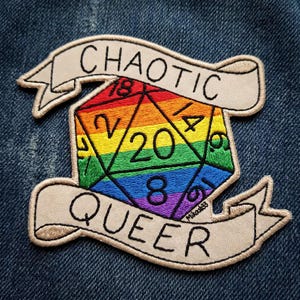 May include: An embroidered patch featuring a rainbow-colored twenty-sided die with black numbers. The die is centered between two beige banners with black stitched text that reads CHAOTIC on the top banner and QUEER on the bottom banner.
