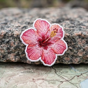 May include: A hibiscus flower patch featuring intricate embroidery in shades of pink, magenta, and deep red. The flower has a prominent yellow stamen extending from the center and is outlined with a clean white border, showcasing detailed stitching.