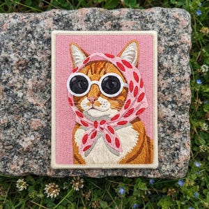 May include: A rectangular embroidered patch featuring an orange tabby cat wearing a pink headscarf with red polka dots and round white-rimmed sunglasses. The cat is set against a solid pink background with a stitched border edge.