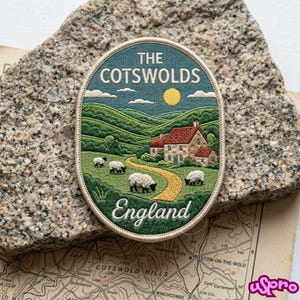 May include: An oval embroidered patch featuring a landscape of The Cotswolds, England. The design shows green rolling hills, a stone house with a red roof, a yellow path, and four white sheep under a yellow sun. The text reads THE COTSWOLDS England.