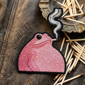 May include: An embroidered patch featuring a pink frog with a black eye, holding a lit cigarette in its mouth. A trail of grey and white smoke rises from the cigarette. The patch has a thick black border and is shaped to follow the frog's outline.