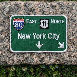 May include: A rectangular embroidered patch with a green background and white border. It features the text East, North, and New York City in white, an Interstate 80 shield, a route 11 shield, and two white arrows pointing downward and right.